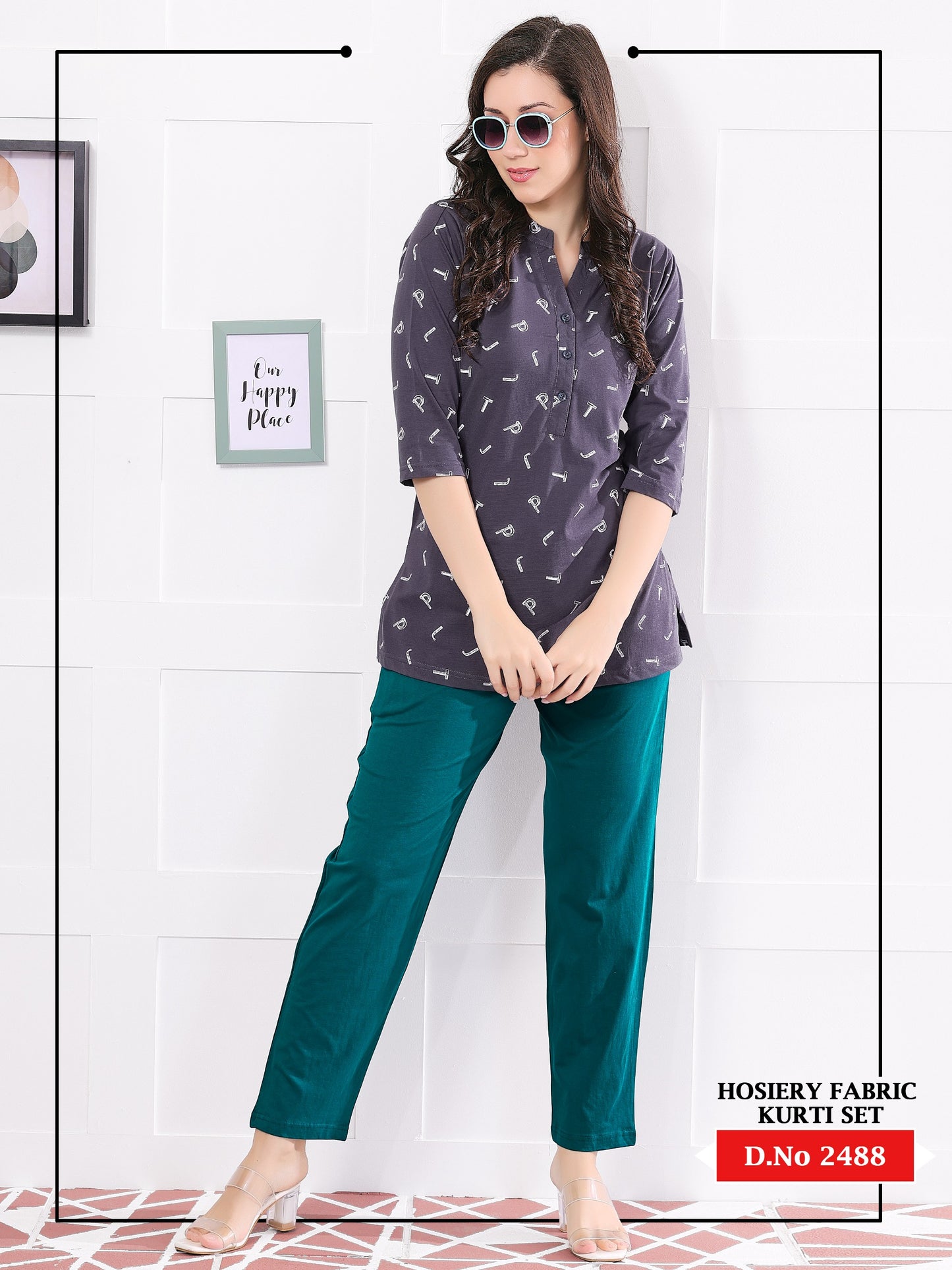 Woman wearing a gray printed top and teal pants against a white tiled wall.