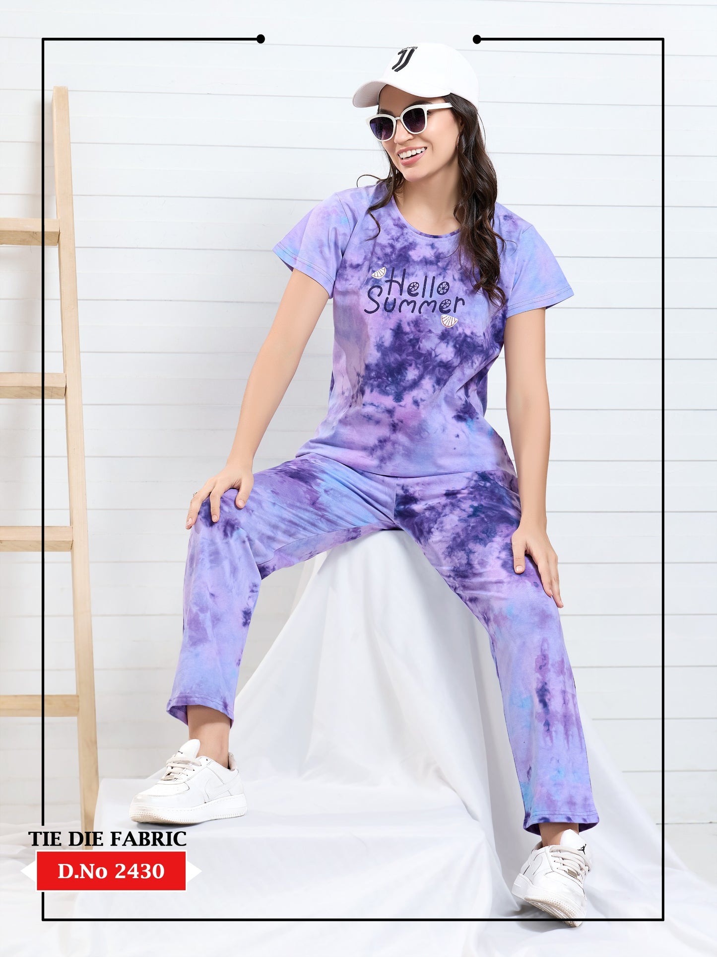 Woman wearing a tie-dye outfit with 'Hello Summer' text, sitting on a white stool against a white wall.