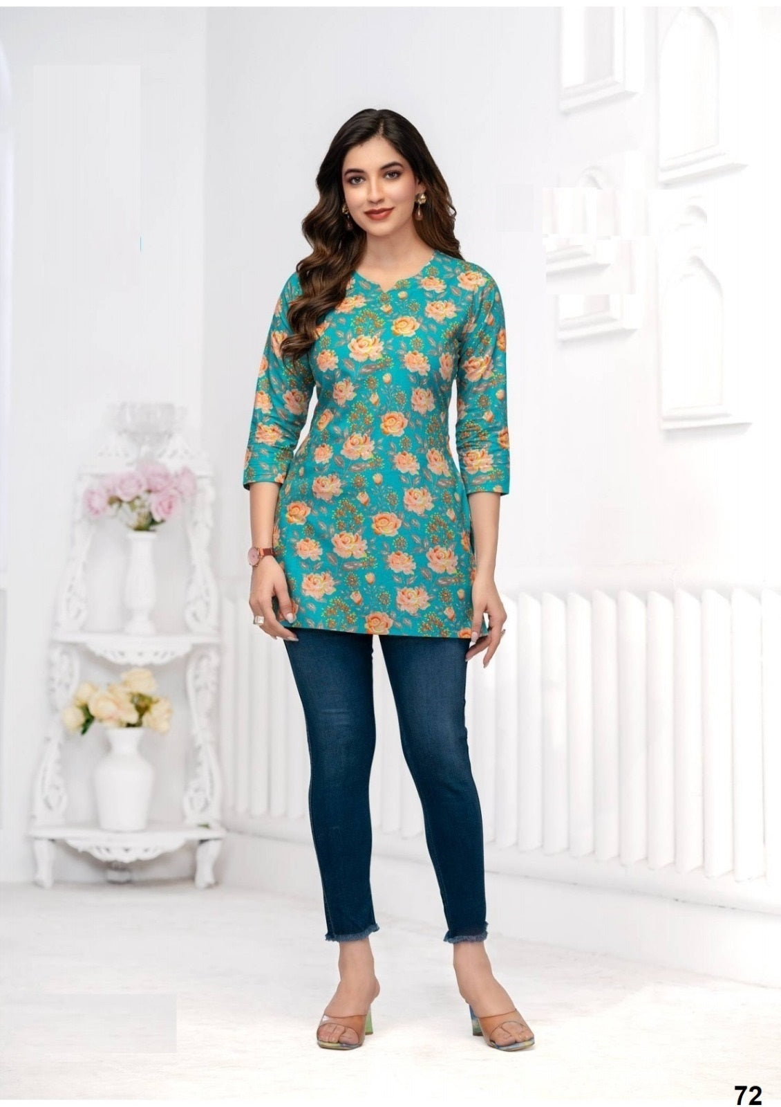 Woman wearing a floral green and blue top with dark blue jeans in a white room.