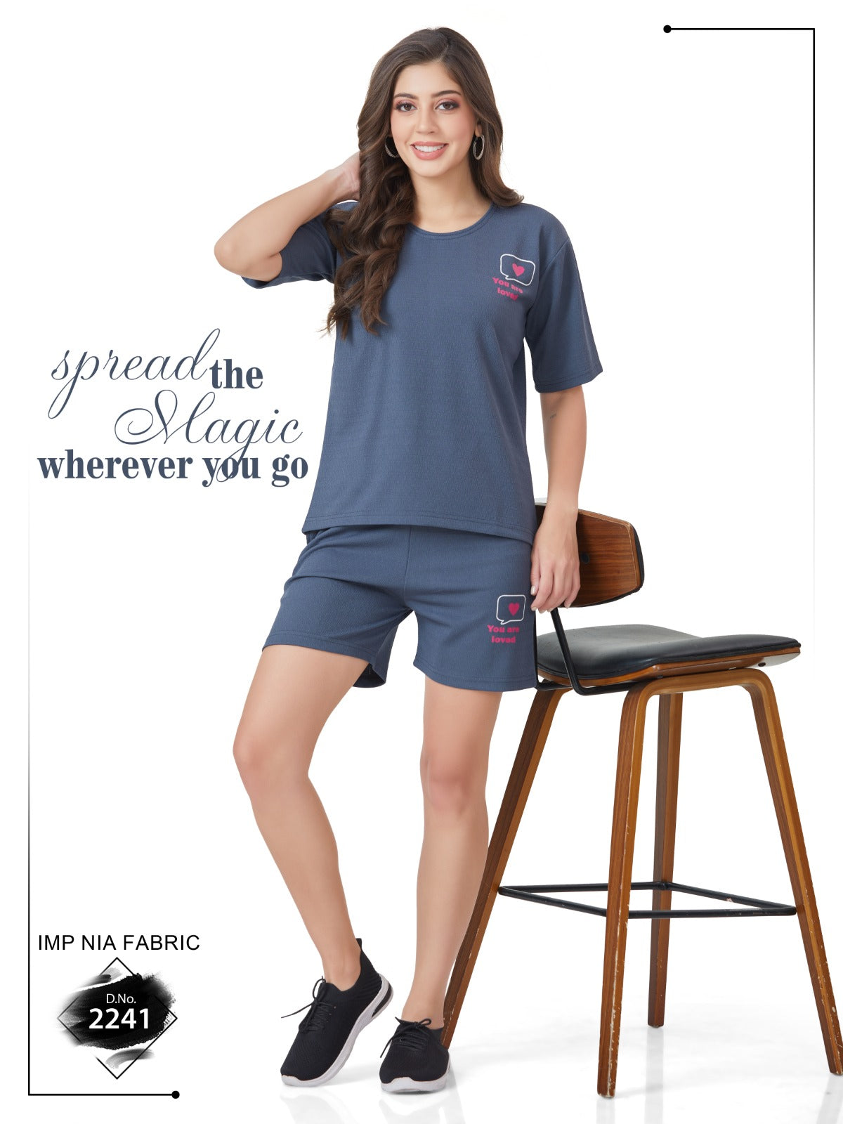 A girl wearing a round neck, short-sleeved blue t-shirt and matching shorts with a graphic design, sitting on a chair next to a wooden table.