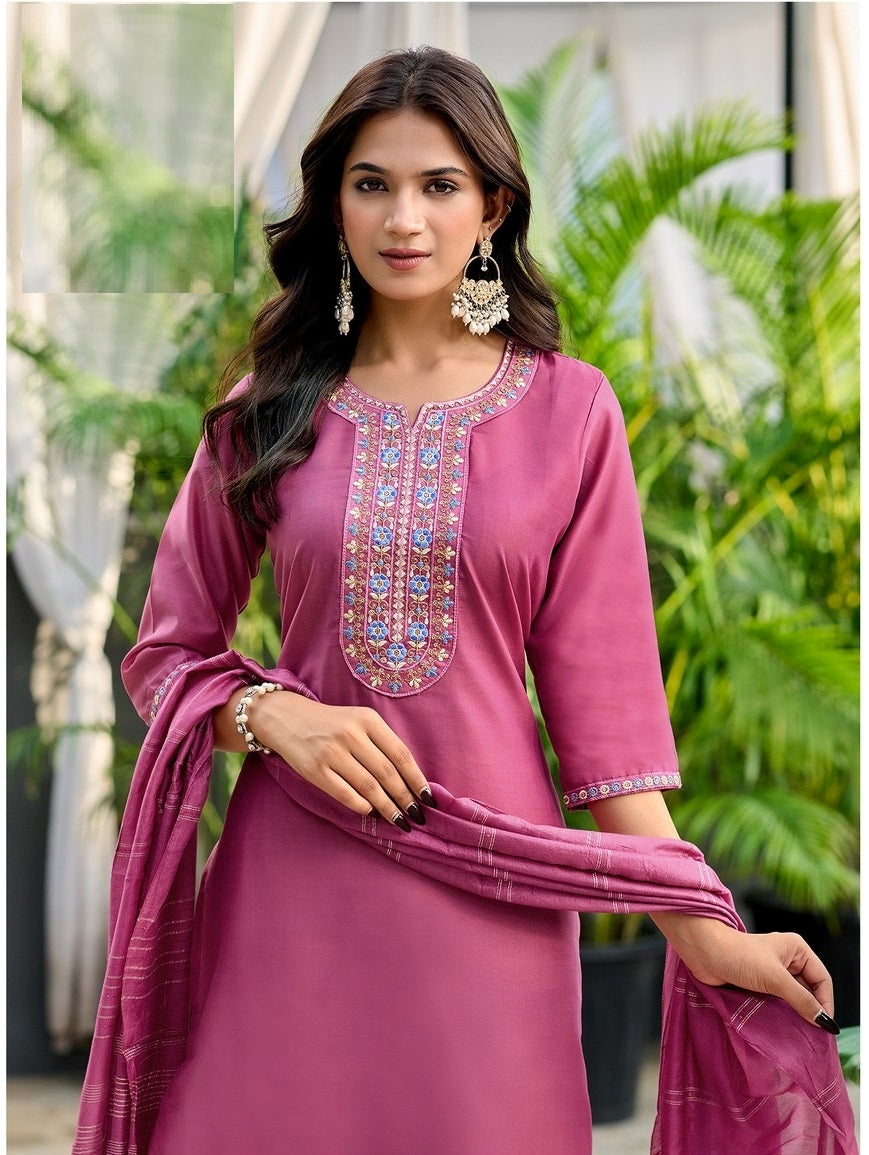 Woman wearing a pink traditional outfit with intricate embroidery, standing outdoors with greenery in the background.