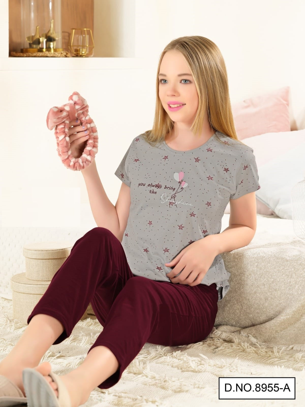A girl sitting on a couch wearing a grey T-shirt with a maroon pants nightsuit. The T-shirt has a cute graphic print and the pants are solid colored.