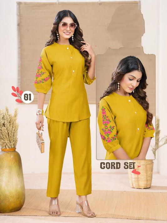 Woman wearing a mustard yellow outfit with floral embroidery, standing against a neutral background.