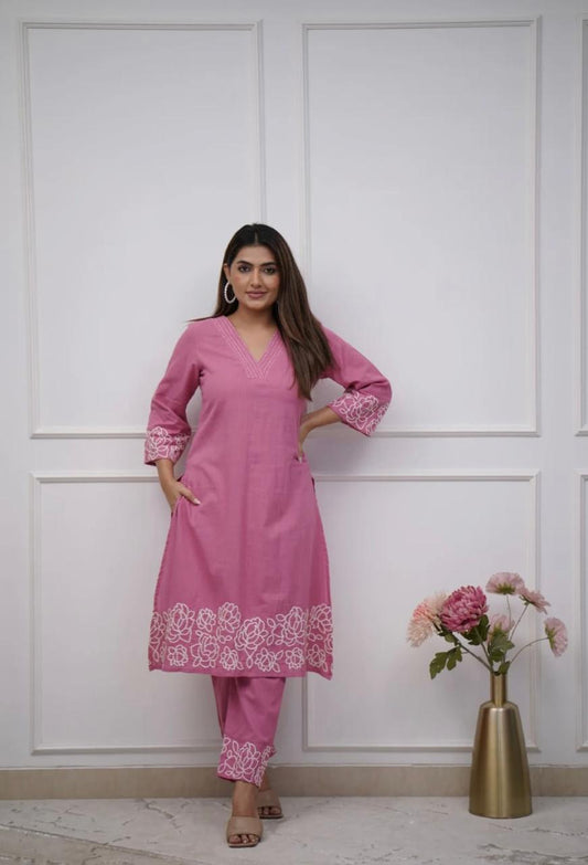 Woman wearing a pink traditional outfit with white embroidery, standing against a white paneled wall.