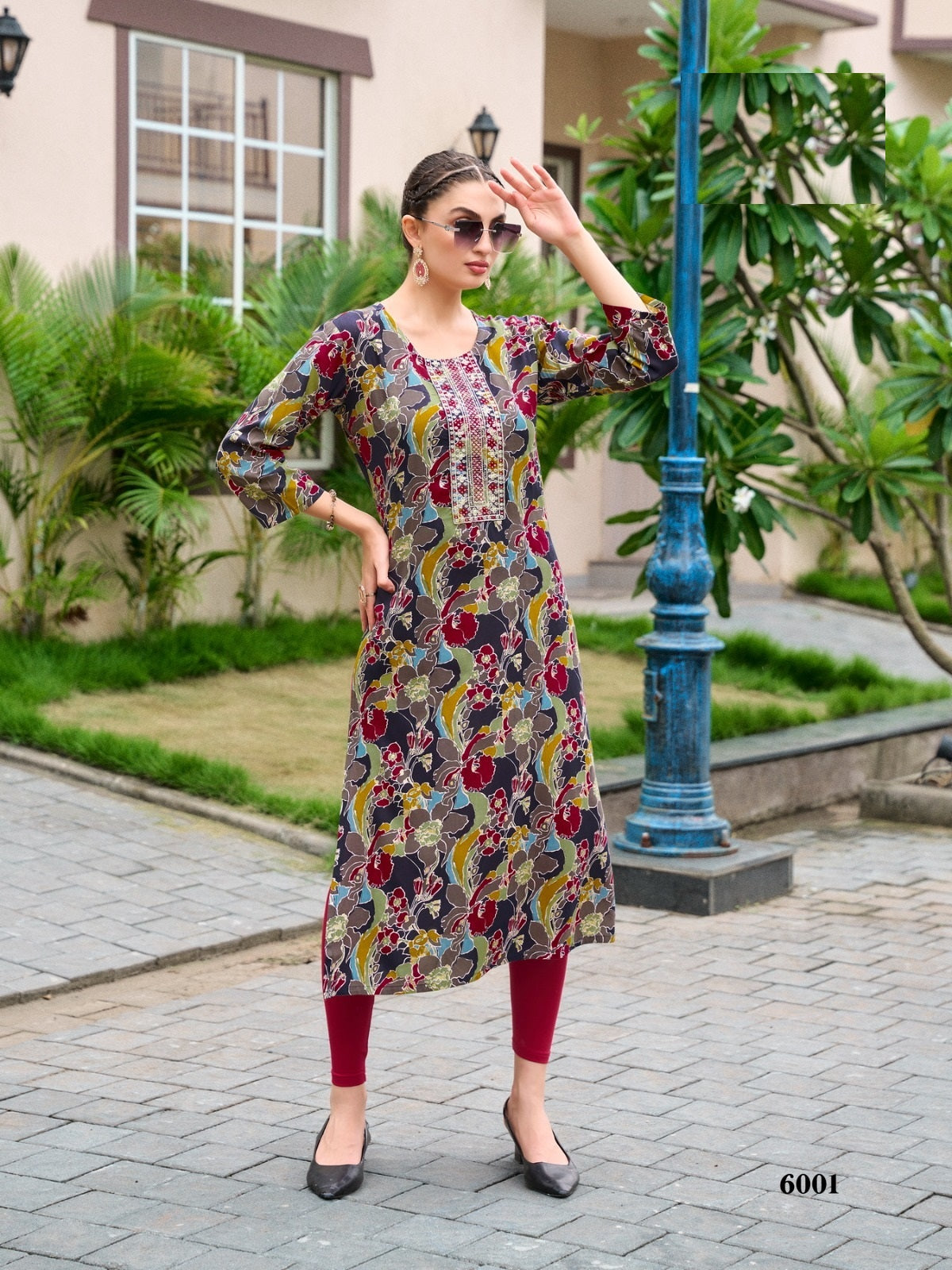 Woman wearing a colorful floral dress in an outdoor setting with greenery and a building in the background.