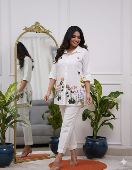 Woman in a white outfit with floral embroidery standing in a room with plants and a mirror.