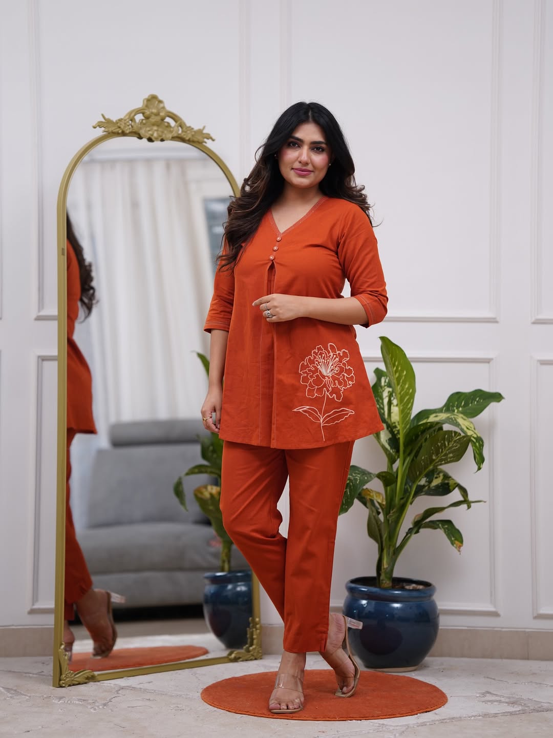 Woman in an orange outfit standing in a room with a mirror and plants.
