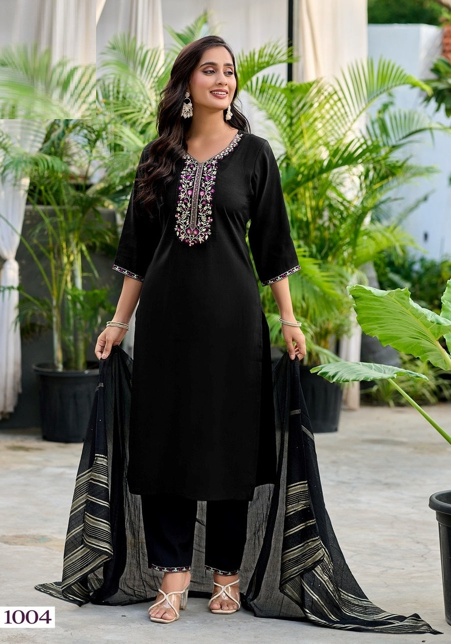 Woman in a black and gray traditional outfit with embroidery, standing outdoors with plants in the background.